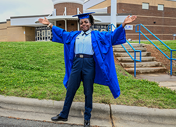  Parkwood High senior with arms extended in front of the school in her JROTC uniform and grad gown.